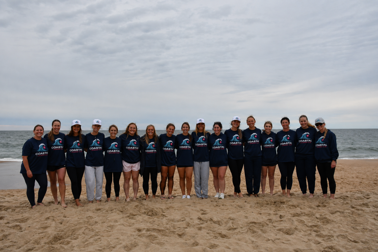 Group of young women standing on the beach in matching coastal team sweatshirts, some wearing caps, smiling at the camera with ocean and cloudy sky in the background.