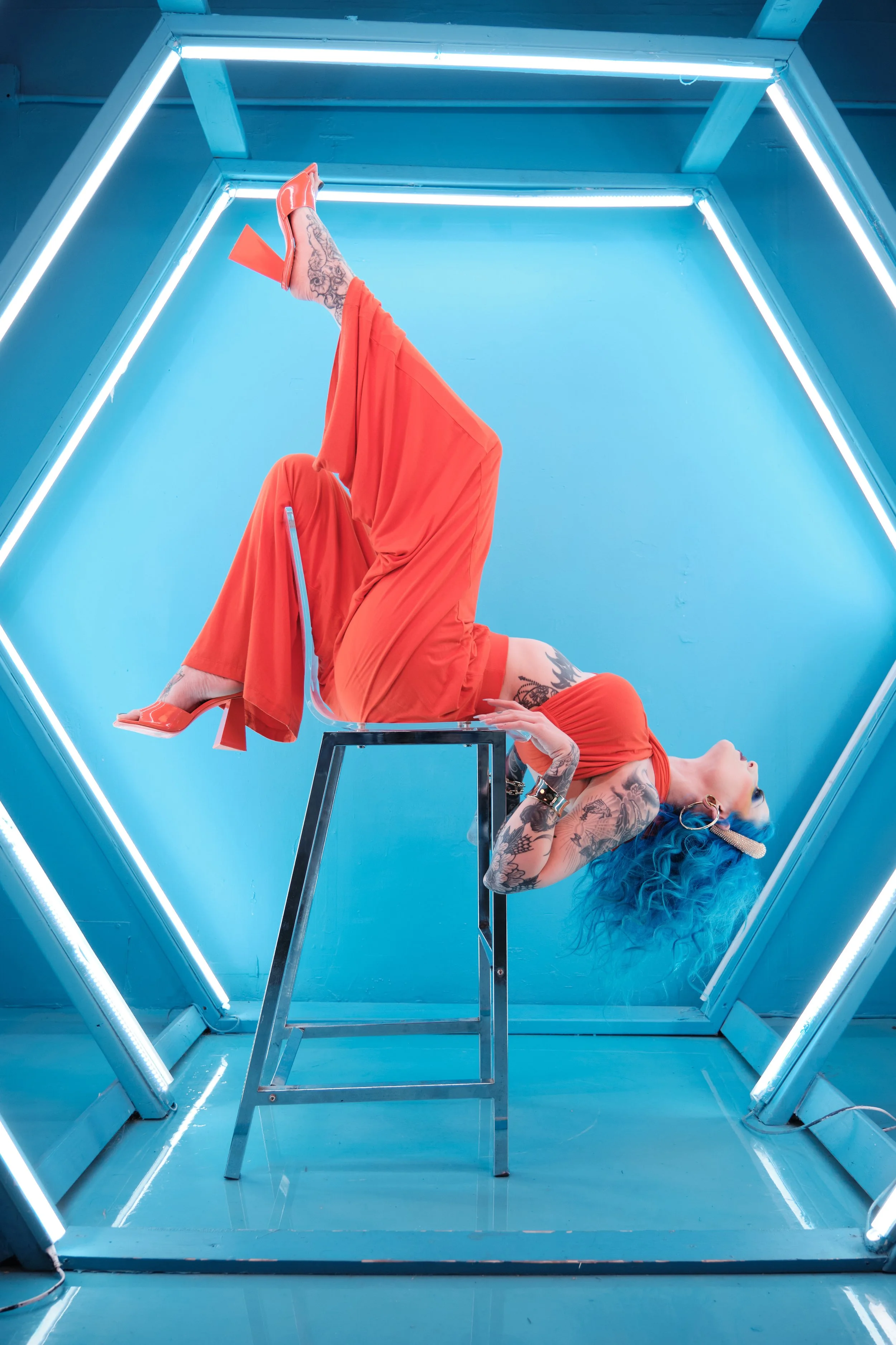 Whitley dressed in an orange outfit and shoes, is lying backwards on a clear chair inside a blue octagonal neon-lit room, with her legs up and feet extended.