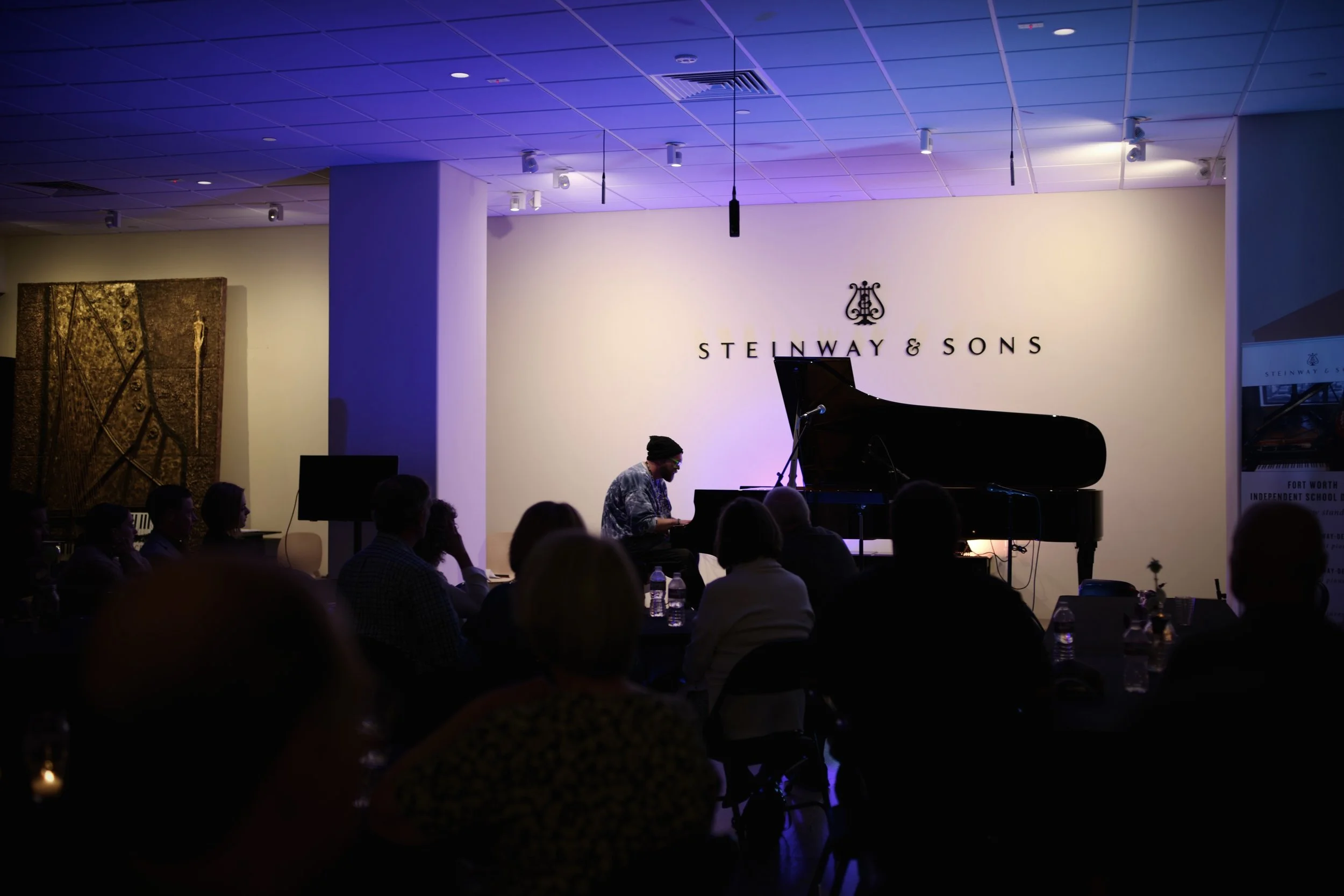 Taylor Collins giving a performance at an event with the sign 'Steinway & Sons' on the wall. Audience members are seated in a dimly lit room with a purple hue from ceiling lights.