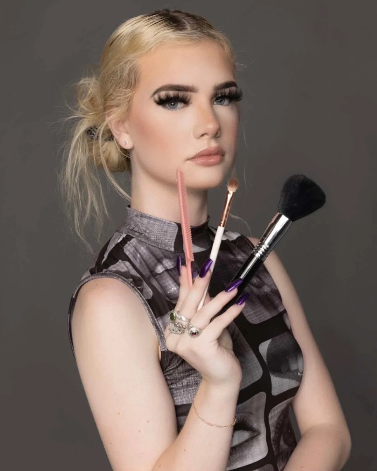 Abby with hair styled in a bun, wearing makeup, earrings, and rings, holding makeup brushes and a comb against a gray background.