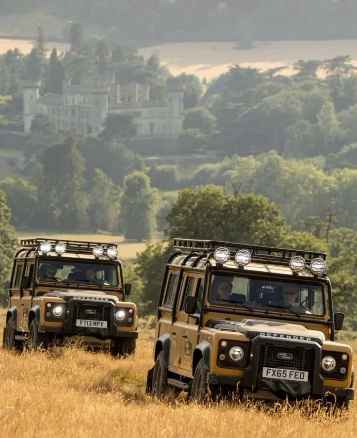 Two beige Land Rover Defenders driving through a grassy field with a castle and trees in the background.