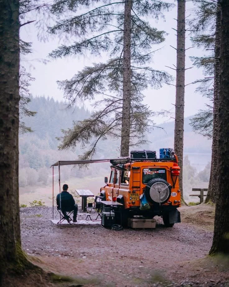 A person sitting in a camping chair with a laptop under an awning, next to an orange off-road vehicle with roof storage, parked in a forest clearing with tall trees and a foggy background.