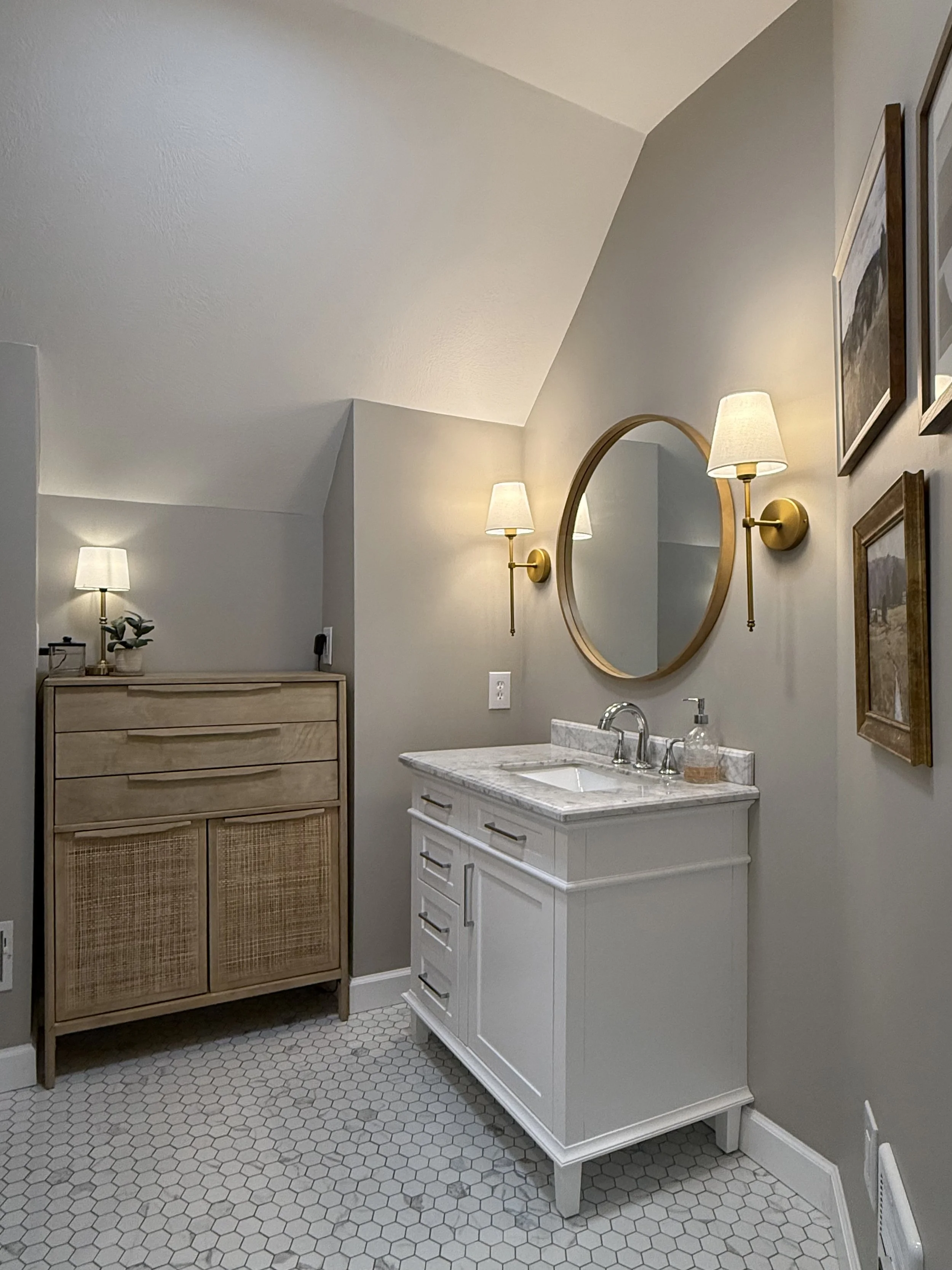A bathroom with white hexagonal tile flooring, a white vanity with a marble countertop and built-in sink, yellow wall-mounted lamps on either side of a round mirror, framed art on the wall, and a wooden cabinet with rattan doors, a small potted plant, and a black device on top.