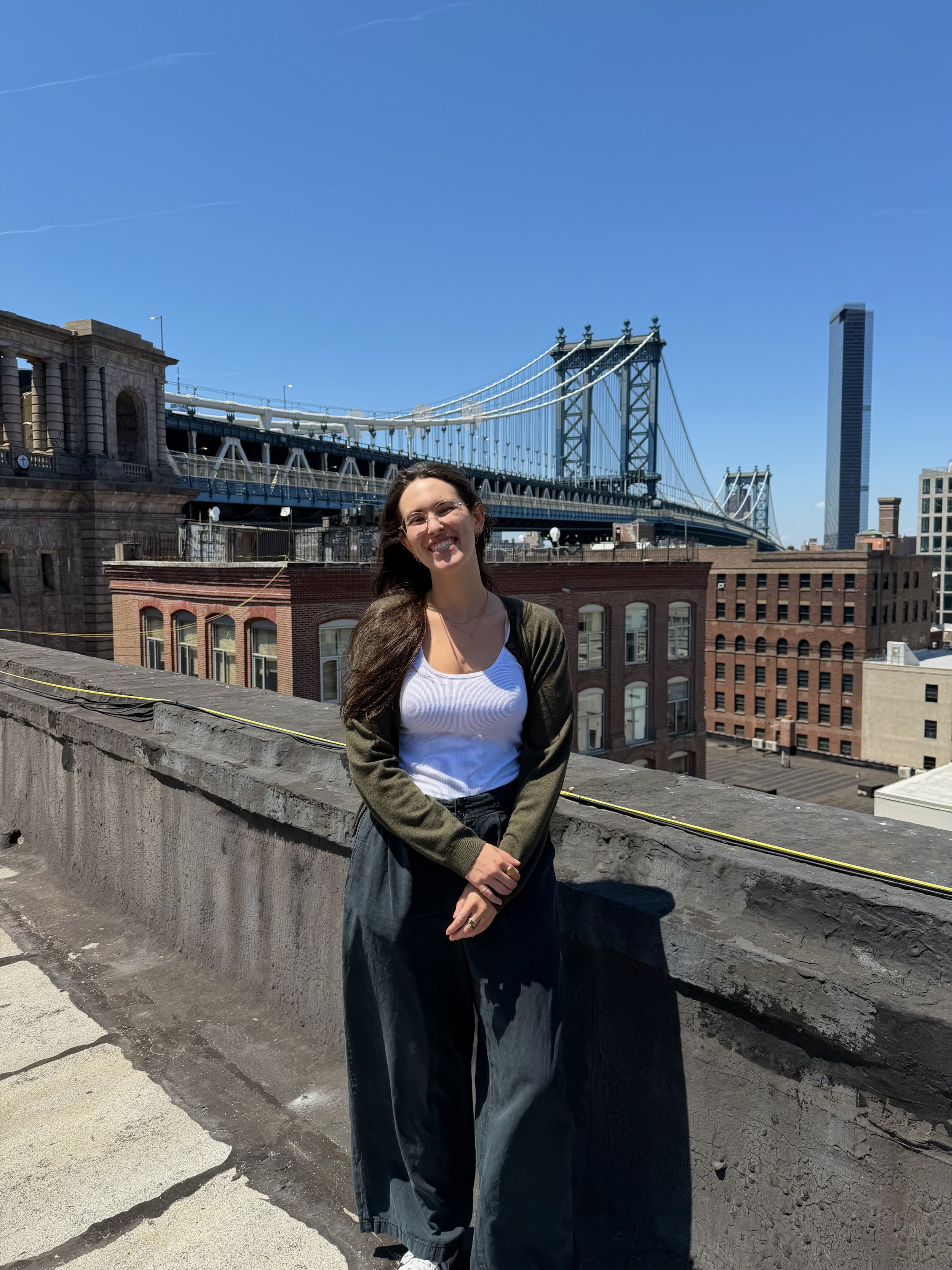 A woman with long brown hair and glasses stands smiling on a rooftop with city buildings and the Brooklyn Bridge in the background under a clear blue sky.