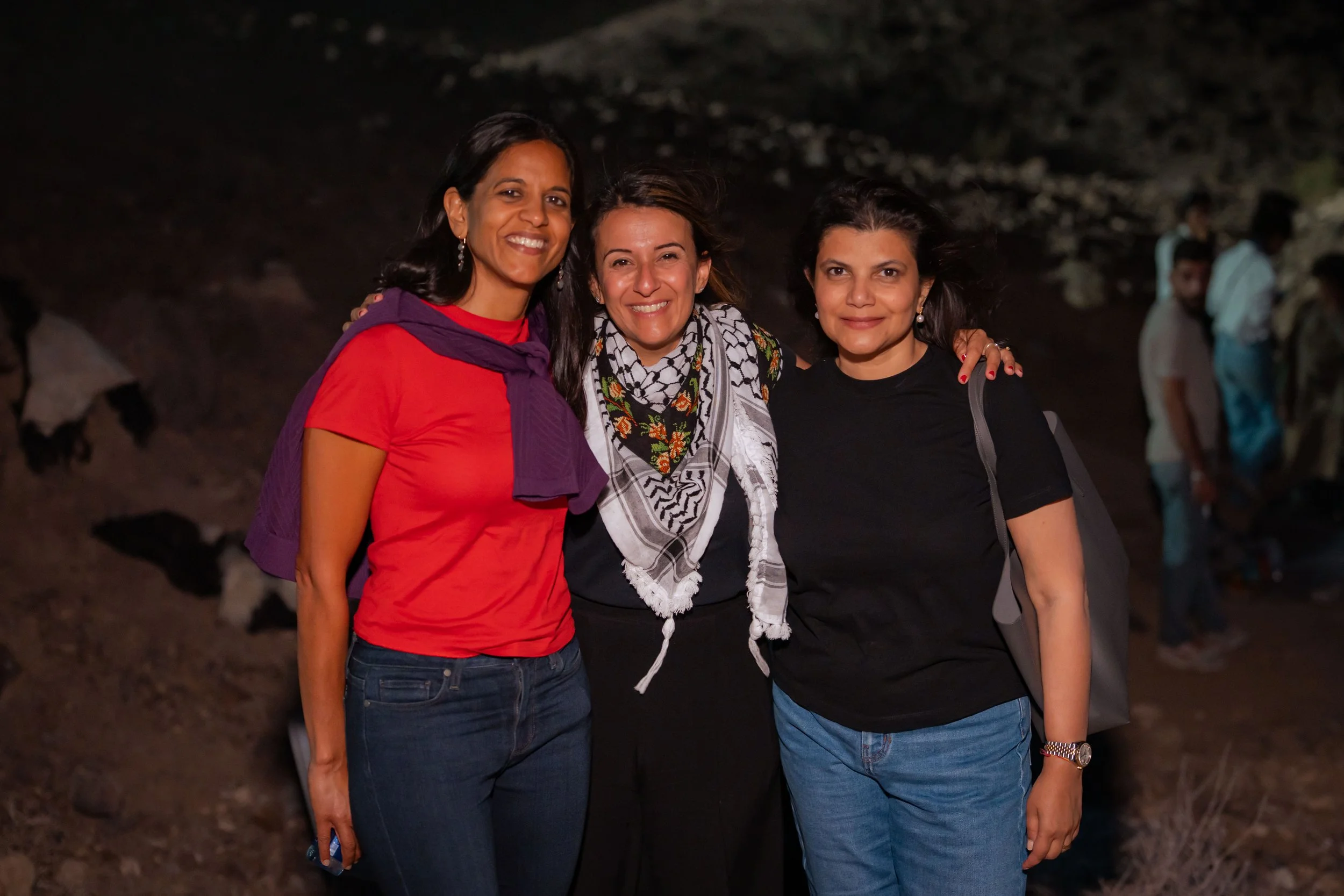 Three smiling women posing together outdoors at night with blurred people in the background.