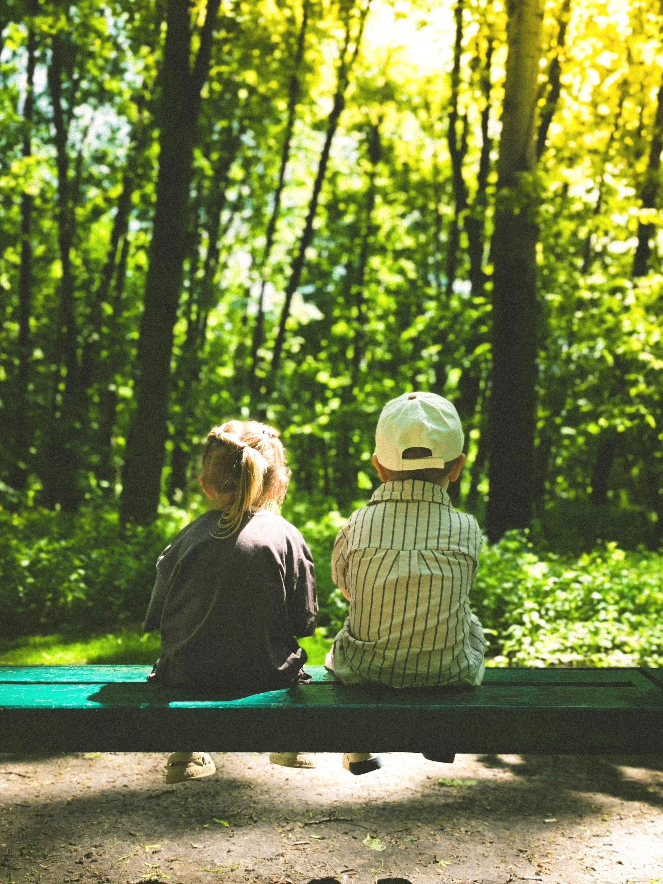 Two children, a girl with braided hair and a boy wearing a white cap, sit on a bench in a green forest, facing away from the camera, during daytime.
