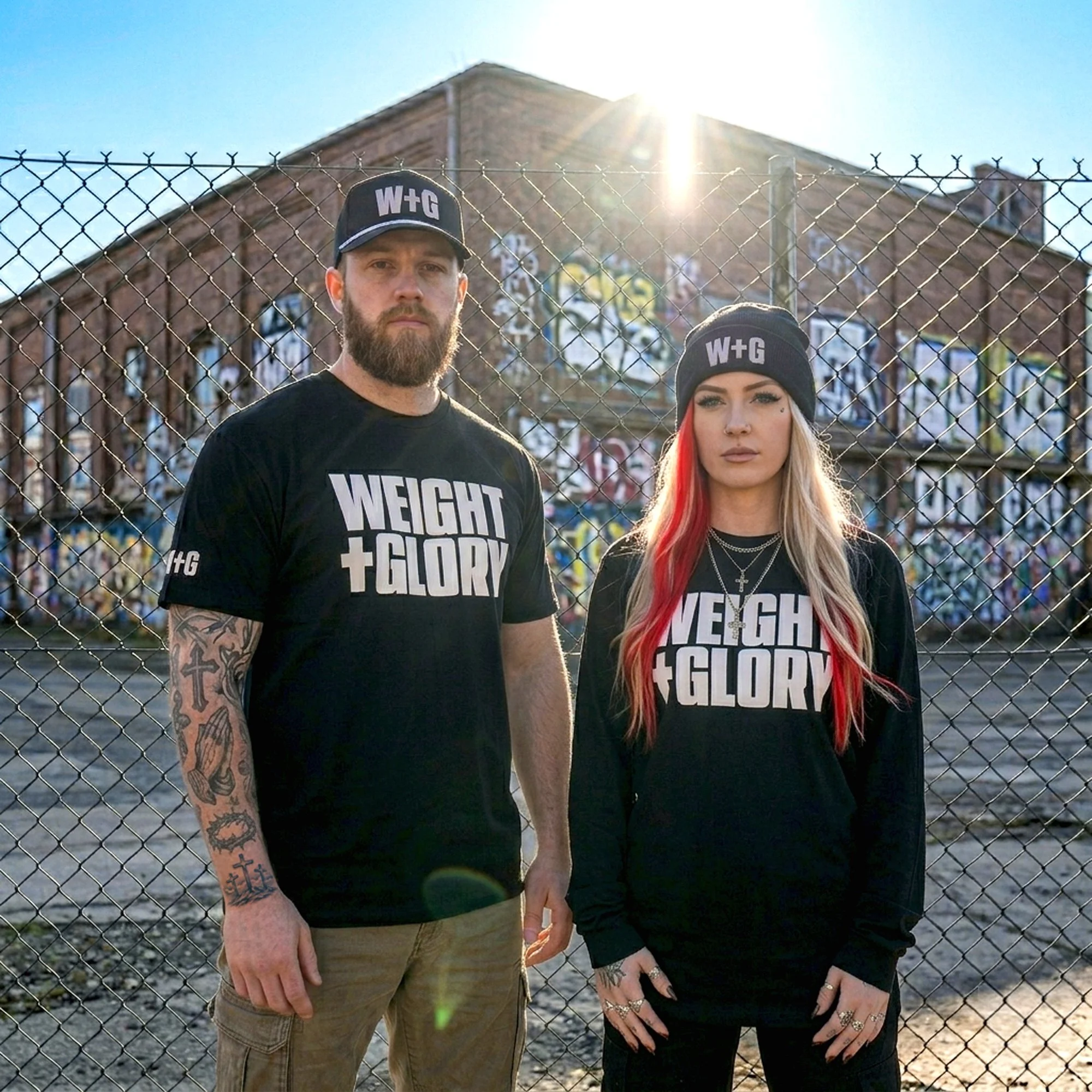 A man and woman standing in front of a chain-link fence with the sun setting behind a brick building. They are both wearing black beanies and black T-shirts with white text reading 'Weight + Glory.' The man has a beard and tattoos on his left arm. The woman has long hair with red streaks, multiple rings on her fingers, and facial tattoos.