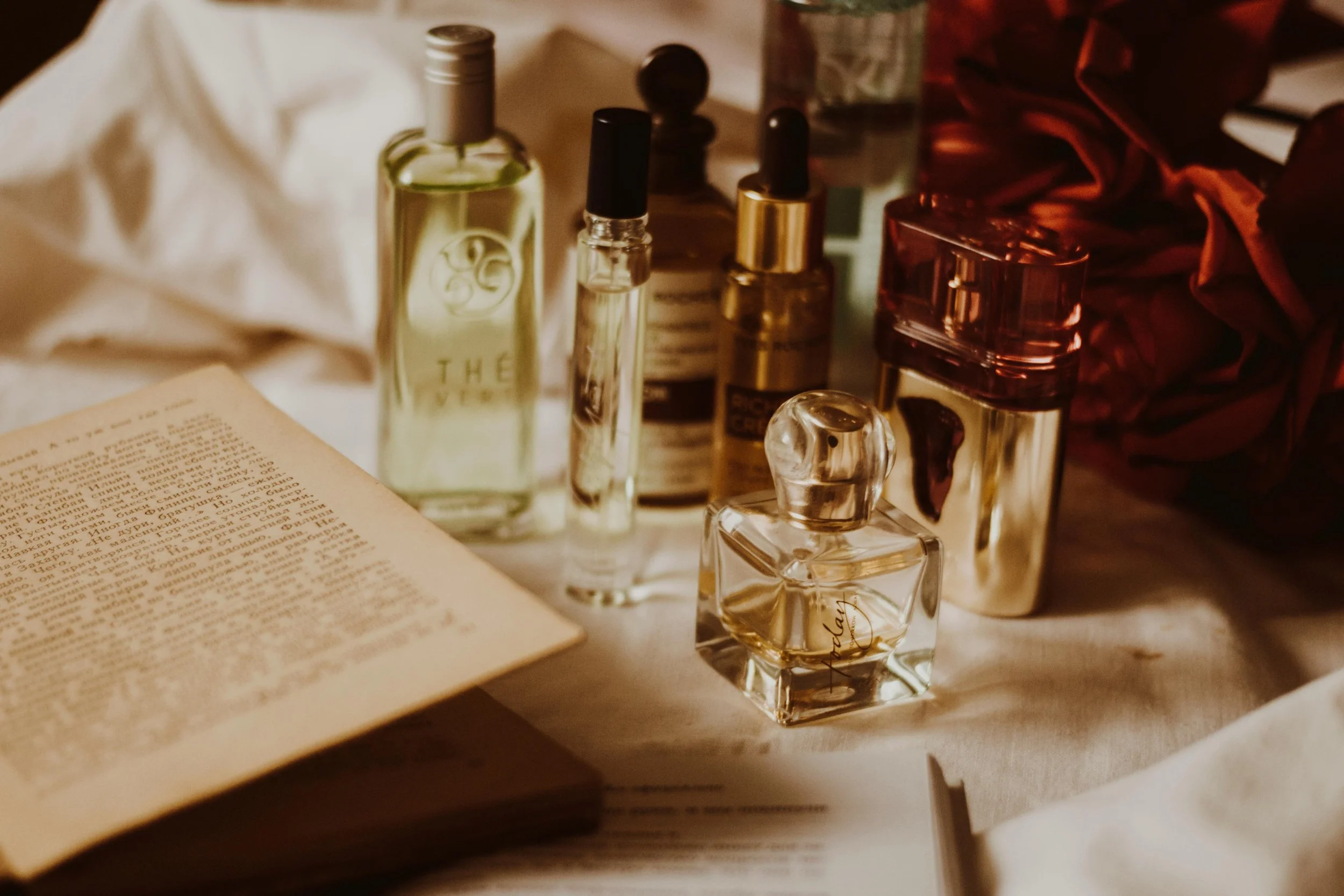 Collection of perfume bottles, an open book, and a bunch of red roses on a white surface.