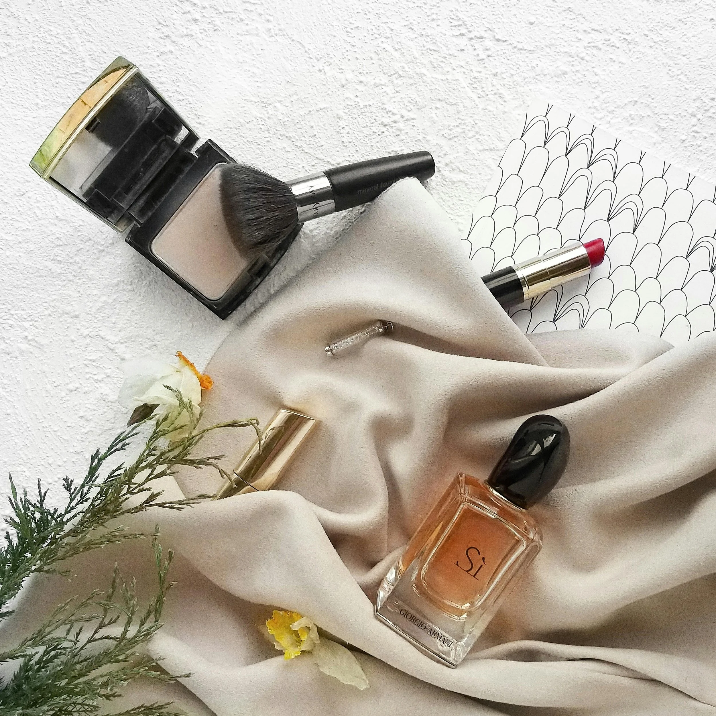 Flat lay of makeup and perfume items on cream-colored fabric, including a makeup brush, compact powder, lipstick, perfume bottle, and decorative paper with a black patterned design, along with flowers and greenery.