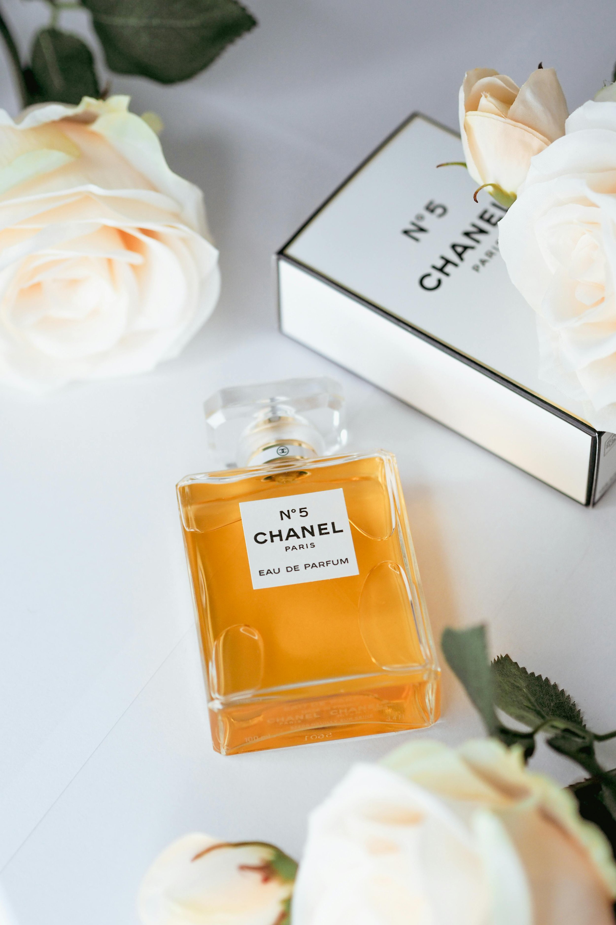 A bottle of Chanel No. 5 perfume next to a box of Chanel No. 5, surrounded by white roses with green leaves, on a white surface.
