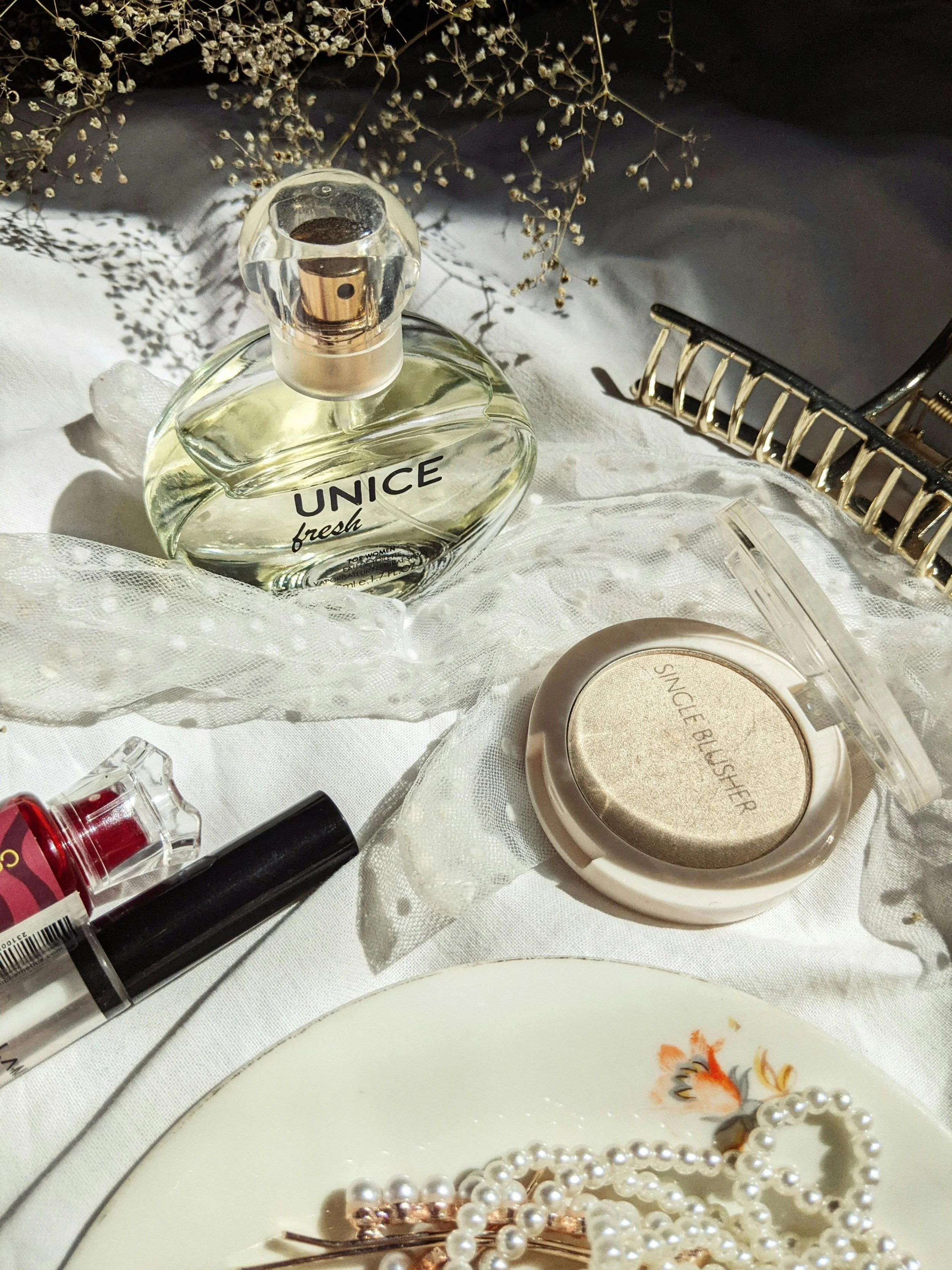 A collection of beauty and accessories items on a white fabric surface, including a bottle of UNICE fresh perfume, a compact single blush, lip gloss, a black eyeliner pen, pearl jewelry, a hair clip, and dried flowers in the background.
