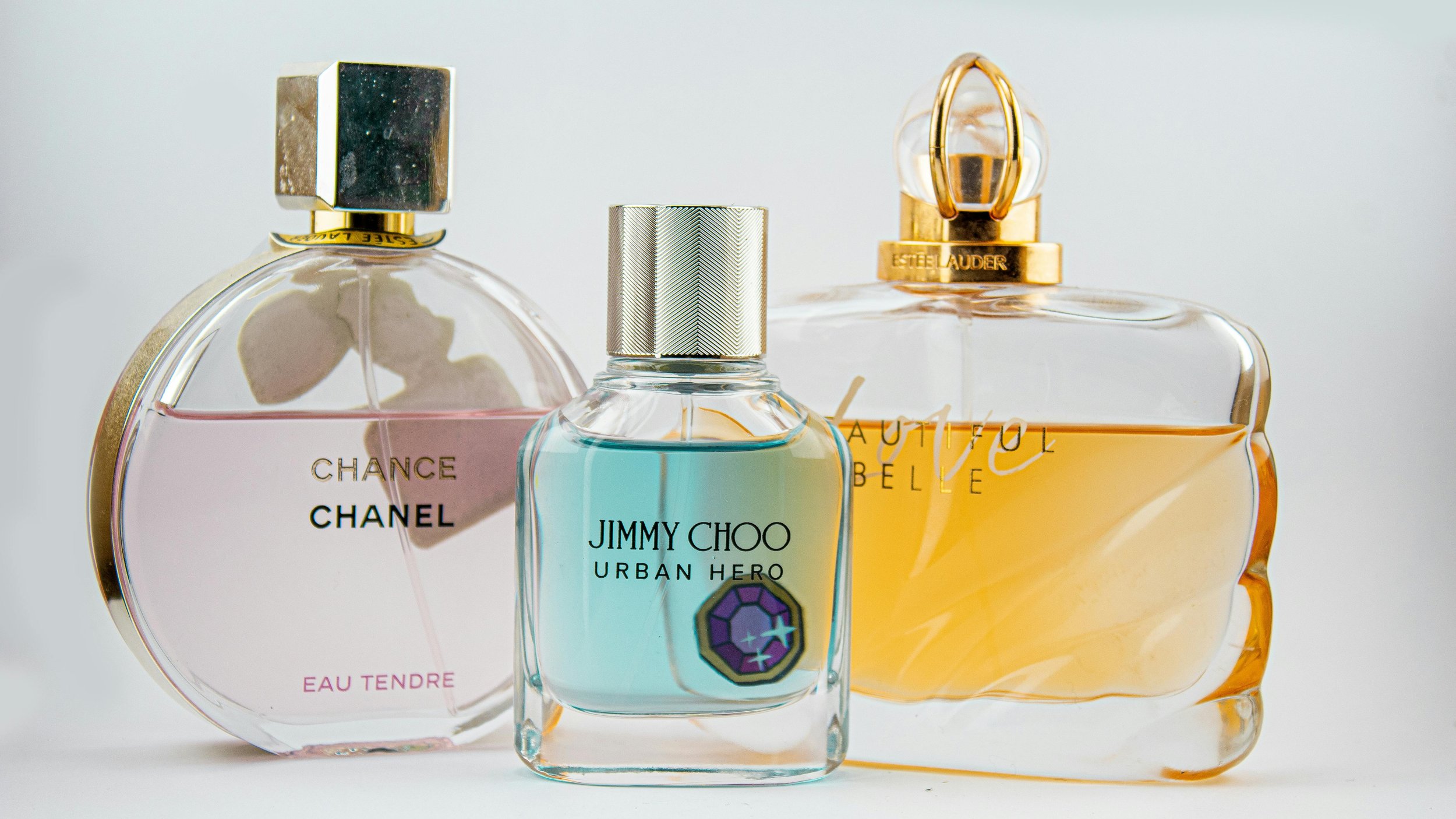 Three perfume bottles, from left to right: Chance Chanel Eau Tendre with a heart design, Jimmy Choo Urban Hero with a blue liquid, and Estée Lauder Beautiful with yellow liquid.