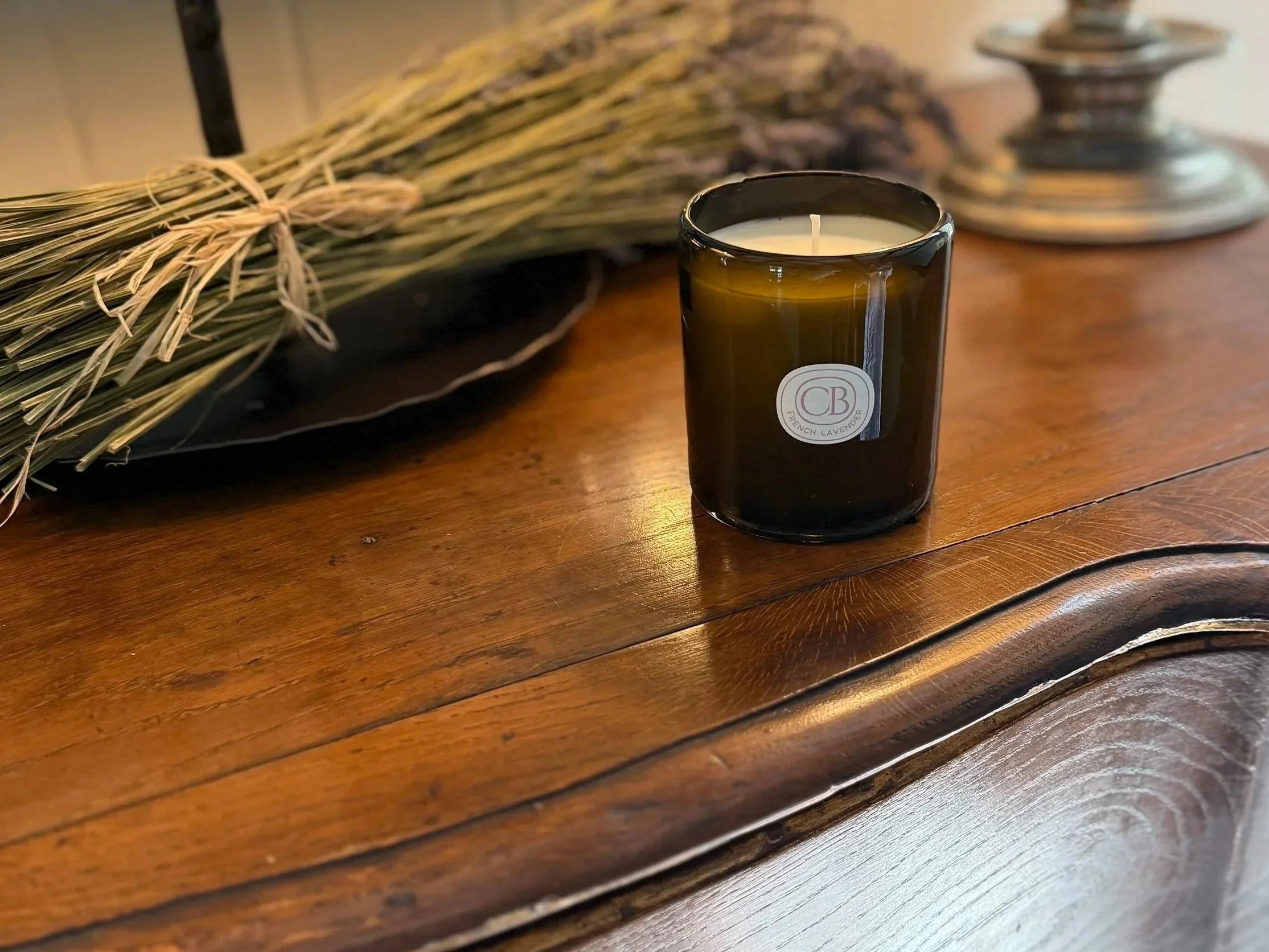A lit Casa Bungo Lavender candle in a jar with a label that says 'French Lavender' on a wooden surface, with a bundle of dried lavender in the background.