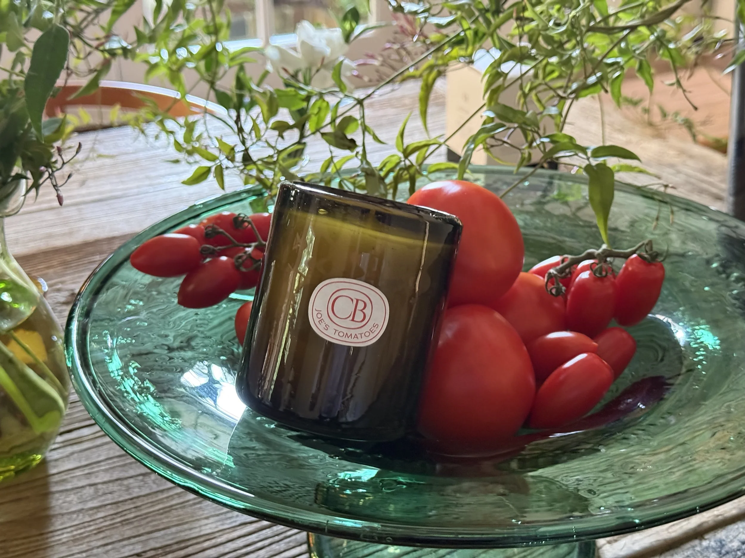 A glass bowl filled with tomatoes and Joe's Tomatoes Casa Bungo candle, with green leaves and branches draping over it, placed on a wooden surface.