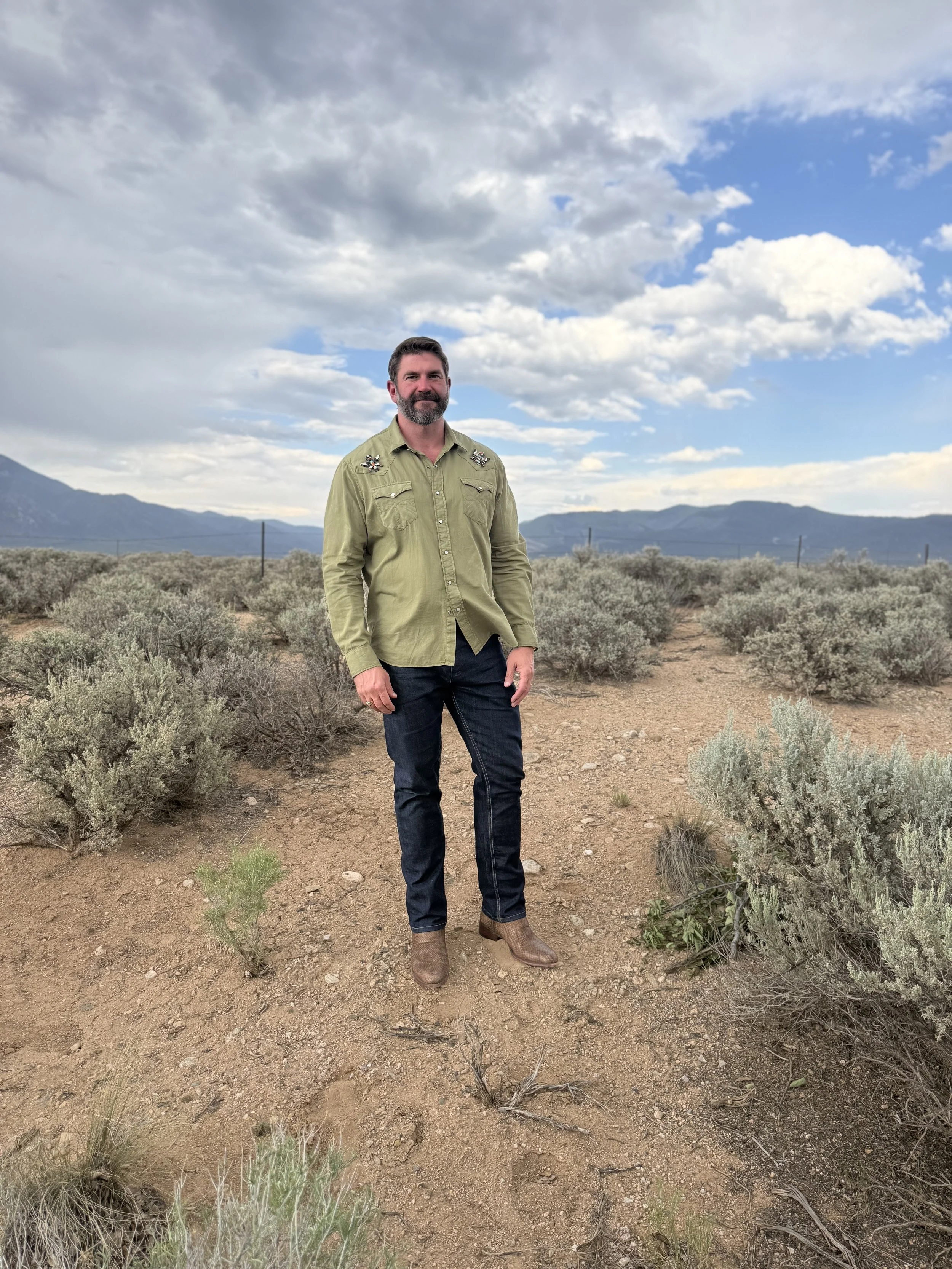A man with a beard and brown hair wearing a khaki shirt with embroidered patches, dark jeans, and brown boots standing in a desert landscape with sparse bushes, mountains in the background, and a partly cloudy sky.
