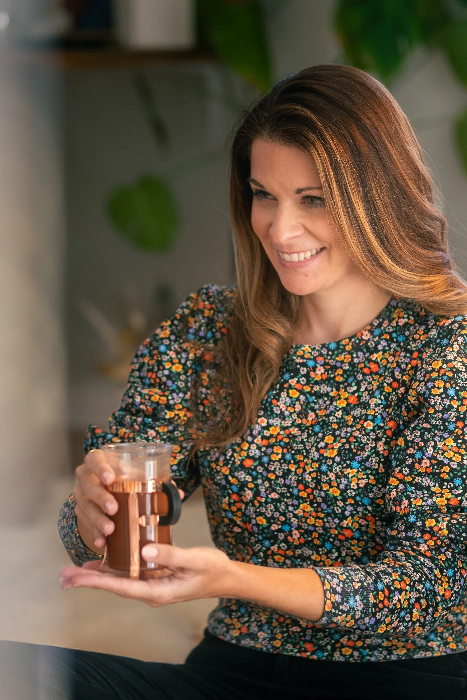 A woman with long brown hair and a floral long sleeve shirt, smiling while holding a copper-colored reusable cup with a black lid, sitting indoors with green plants in the background.