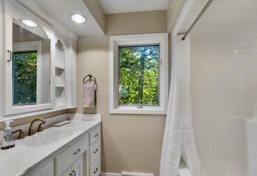 Bathroom with beige walls, white vanity with gold fixtures, a mirror with built-in shelving, a window with green trees outside, and a shower with a white curtain.