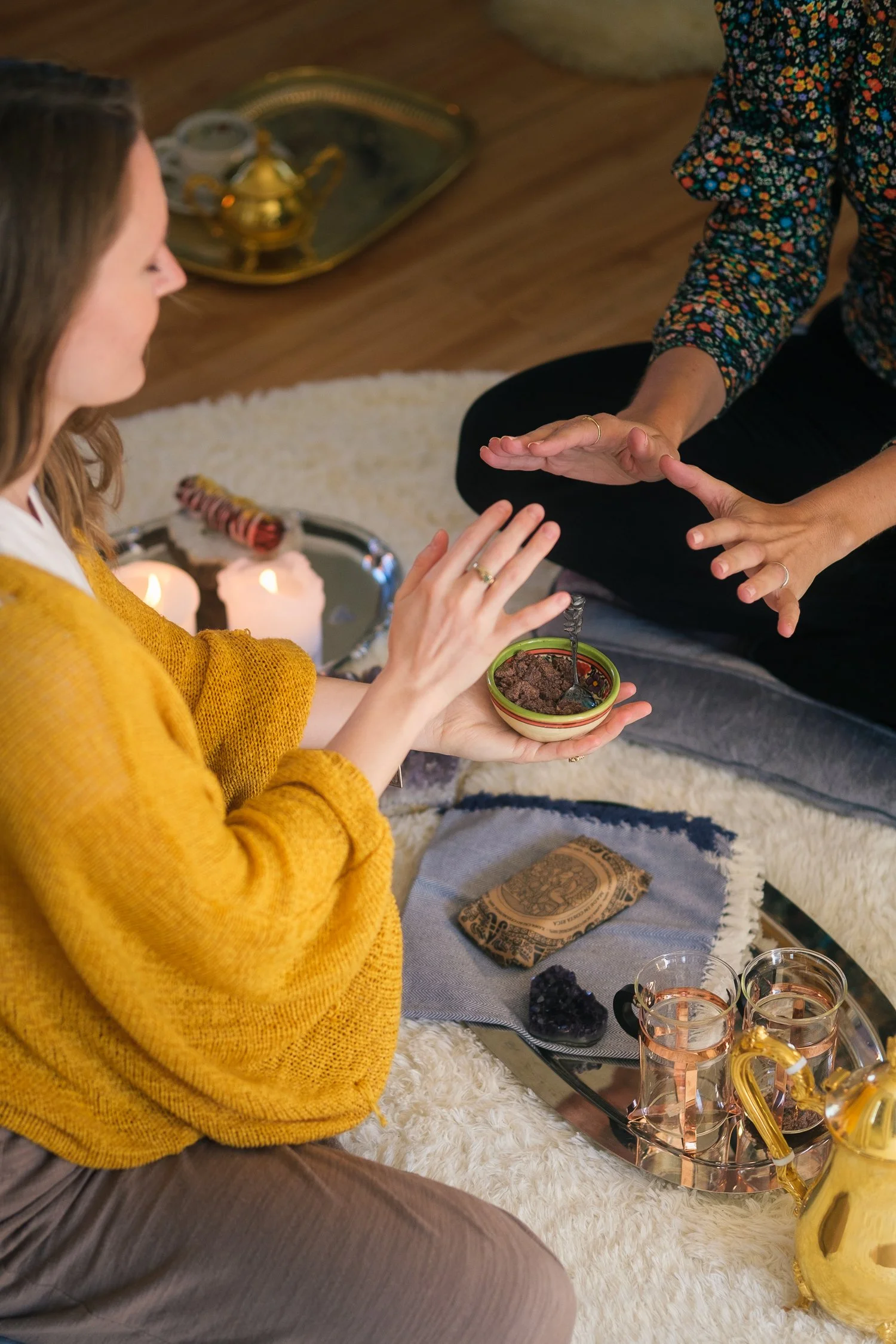 Two women are sitting on a white fuzzy rug, sharing a dessert. One woman is wearing a mustard yellow sweater, and the other is wearing a floral top. There is a tray with glasses, a yellow pitcher, and a bowl of dessert between them, along with candles and decorative items on the tray and nearby.