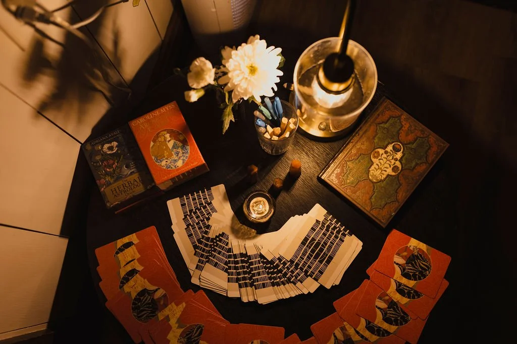 A table with tarot cards fanned out, tarot card decks, a lit candle, a glass of water with a straw, an open book with a decorative cover, a small jar with matches, and a flower in a vase, all under warm lighting.