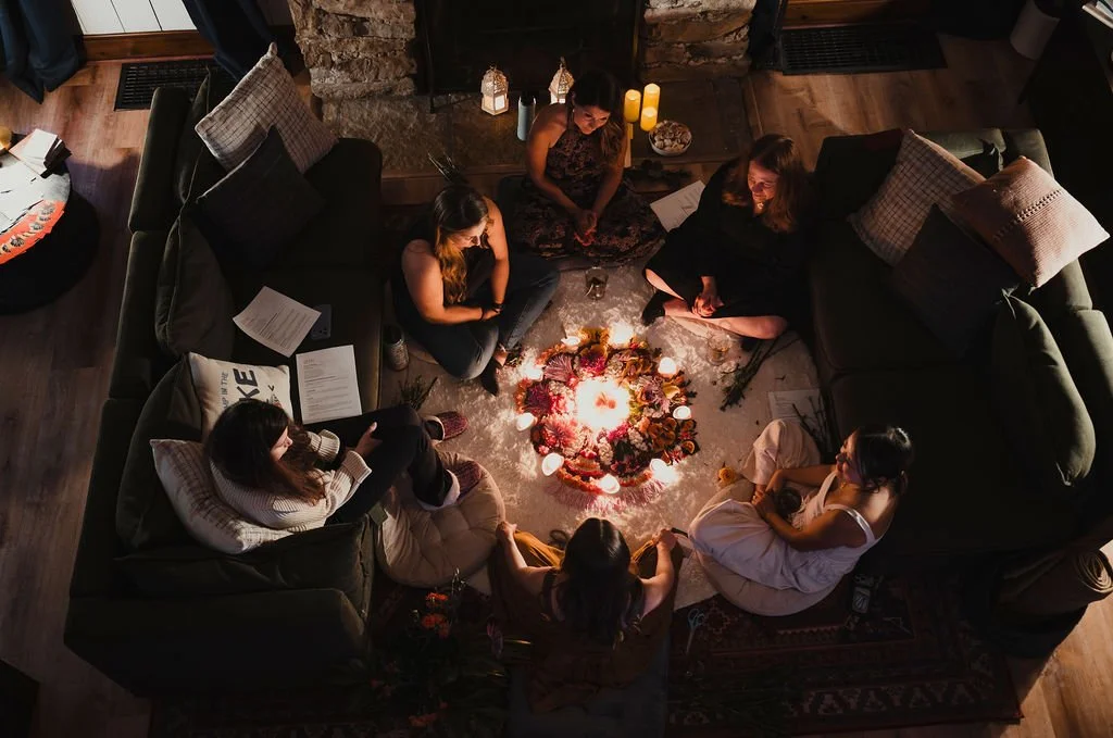 Six women sitting on couches and cushions in a cozy living room, gathered around a lit candle centerpiece on the floor, with some books and papers nearby, enjoying a relaxing evening.