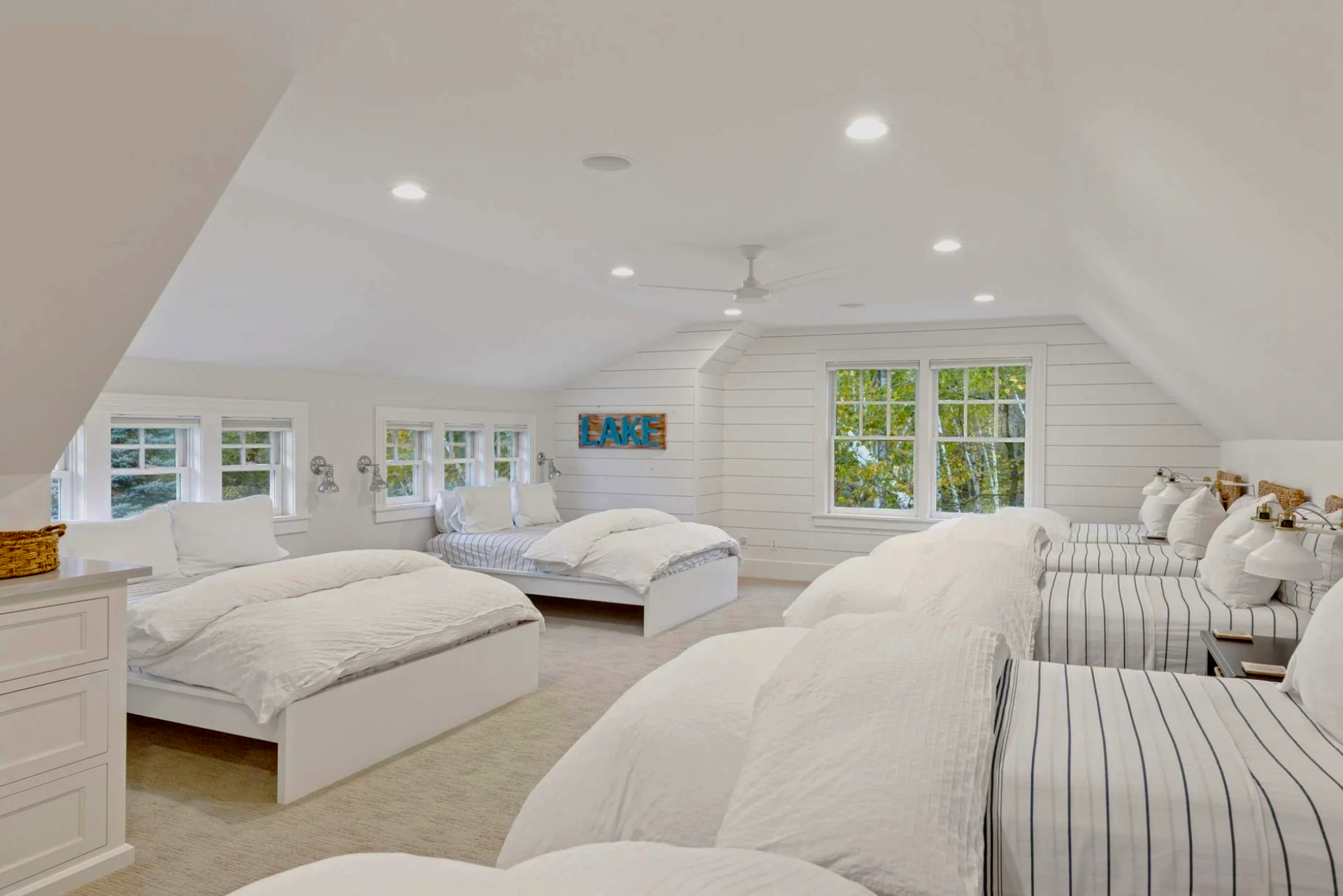 Bright, spacious bedroom with multiple white beds, striped and plain white bedding, and large windows showing green trees outside. There's a sign reading 'LAKE' on the wall and a ceiling fan.