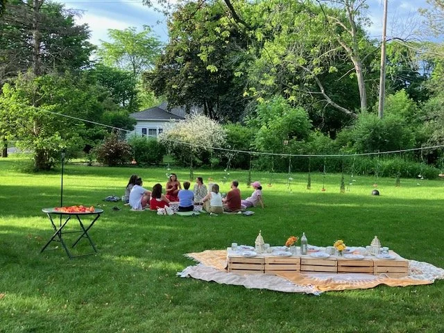 A group of people sitting in a circle on the grass in a backyard, with a picnic setup nearby, under a canopy of trees on a sunny day.