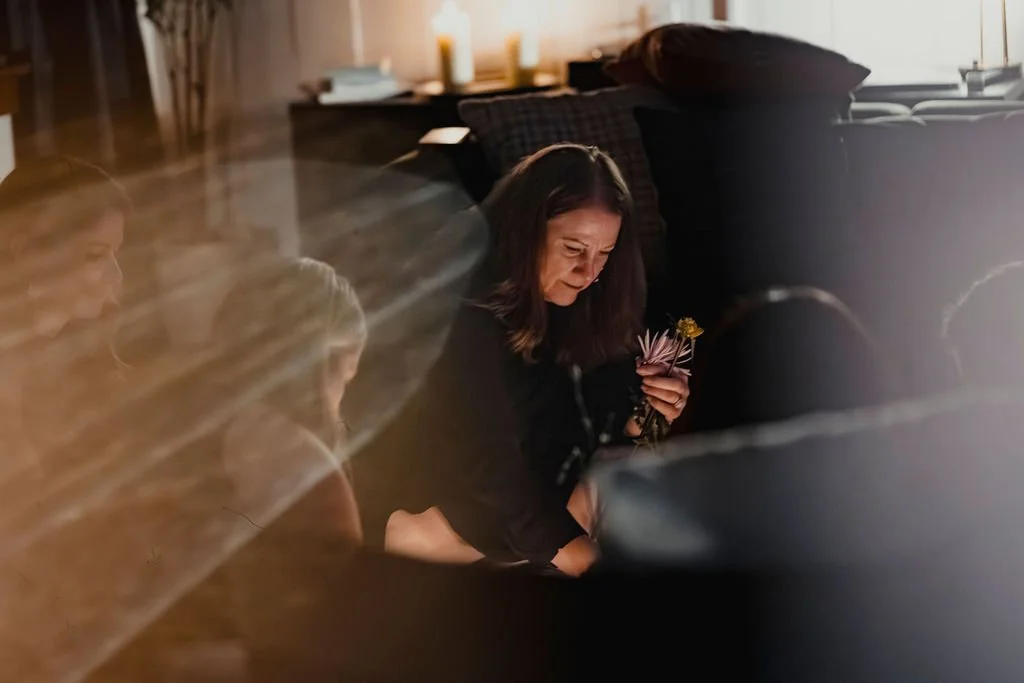 A woman sitting on the floor beside a couch, holding flowers, with a somber expression in a cozy living room with candles.
