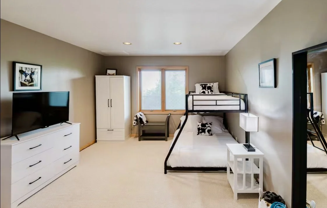 Bedroom with a bunk bed, a dresser with a TV, a white wardrobe, a window, and a chair with a blanket.