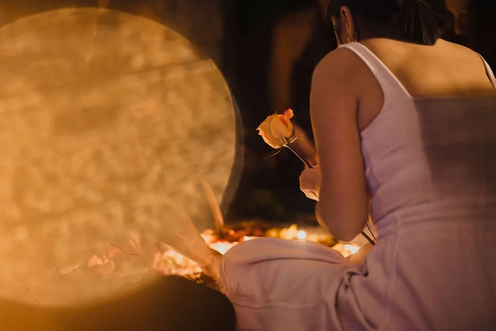 A woman in a white tank top holding a yellow rose at night, with the scene illuminated by warm, low lighting.