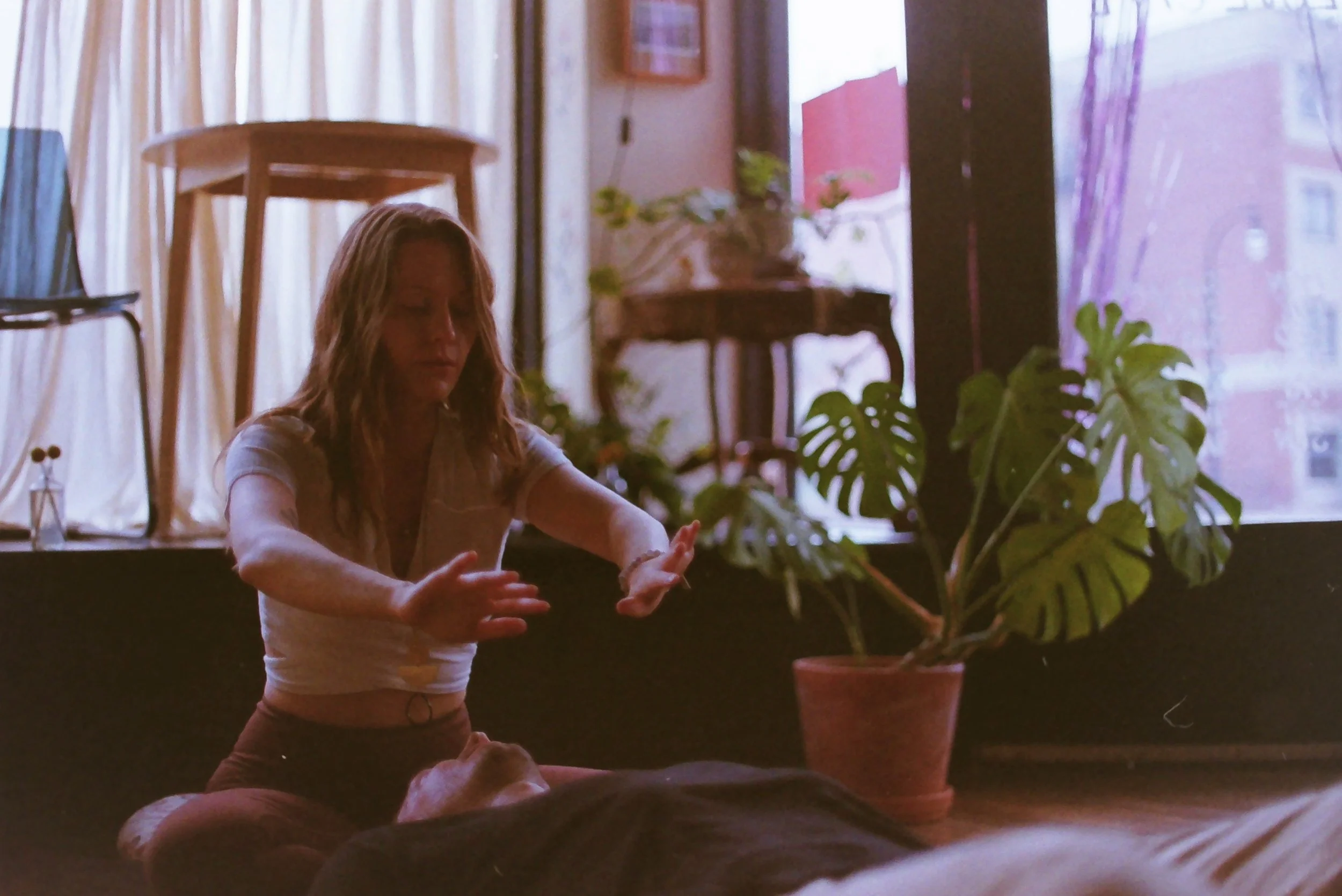 A woman practicing yoga or meditation indoors with plants and sunlight streaming through curtains.