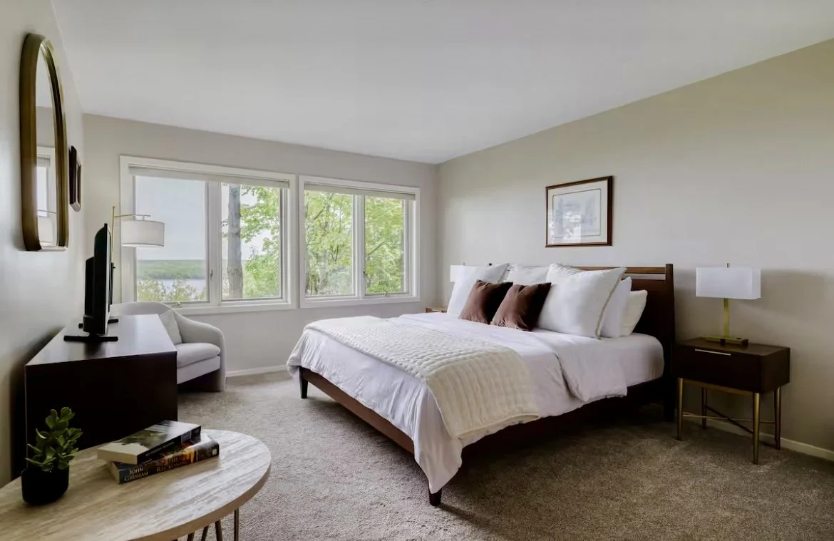 Bedroom with large window, white bed with pillows, side table with lamp, dresser with TV, small table with books and plant.
