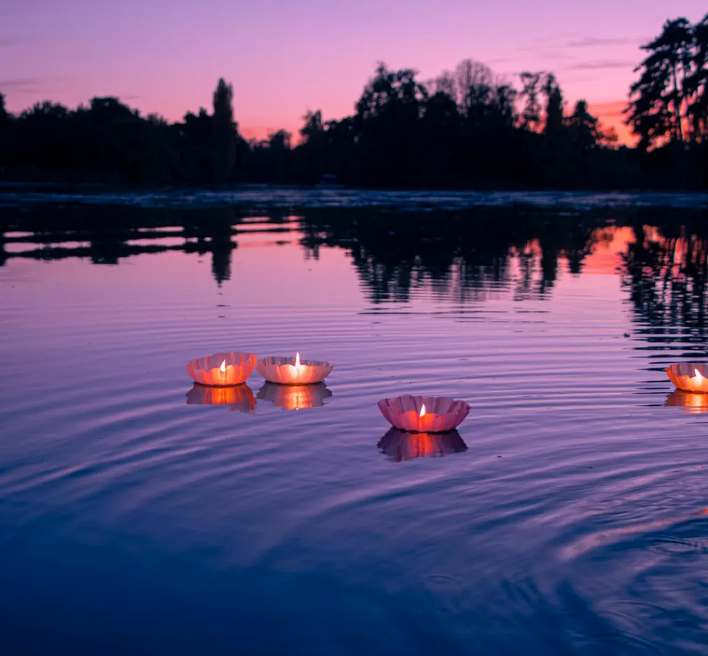 Four floating paper lanterns on a calm river at sunset with pink and purple sky and silhouetted trees in the background.