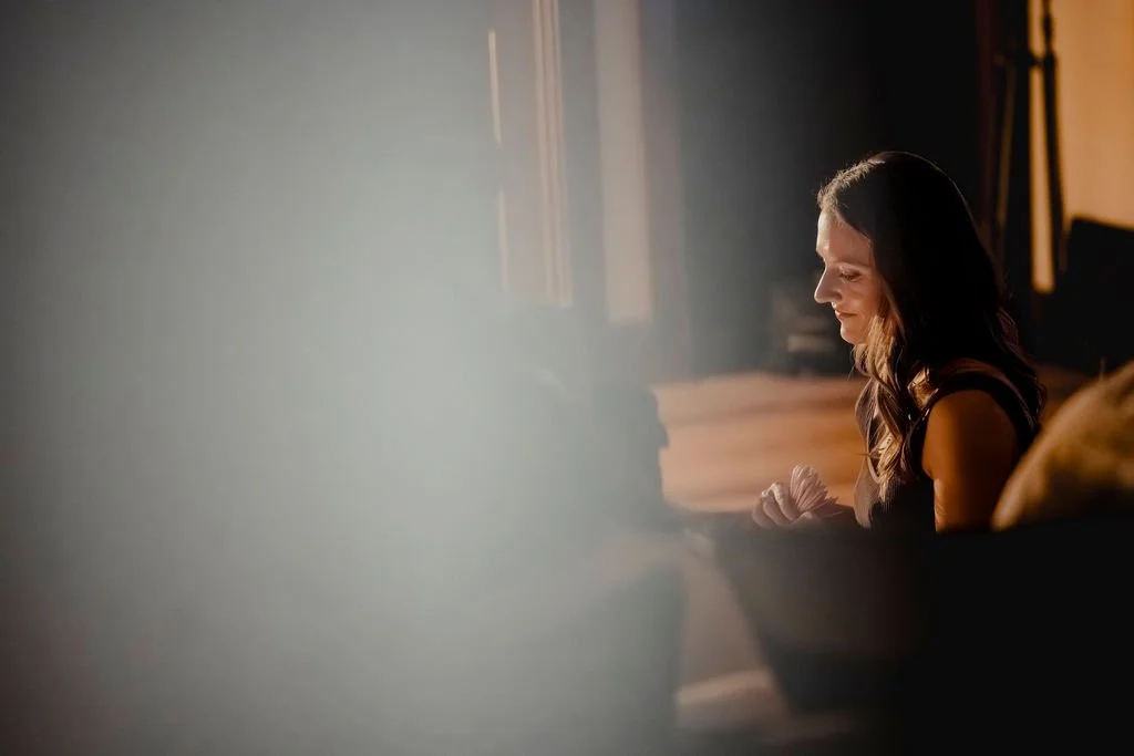 A woman sitting on a sofa in a softly lit room, with sunlight coming through a window, appears to be in a moment of reflection or prayer.