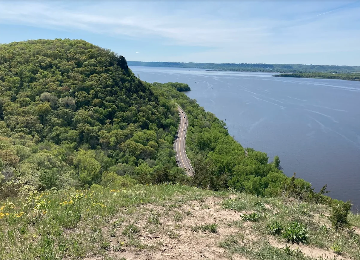 A scenic view of a river bordered by green trees and a winding road on a hill, with a partly cloudy sky overhead.