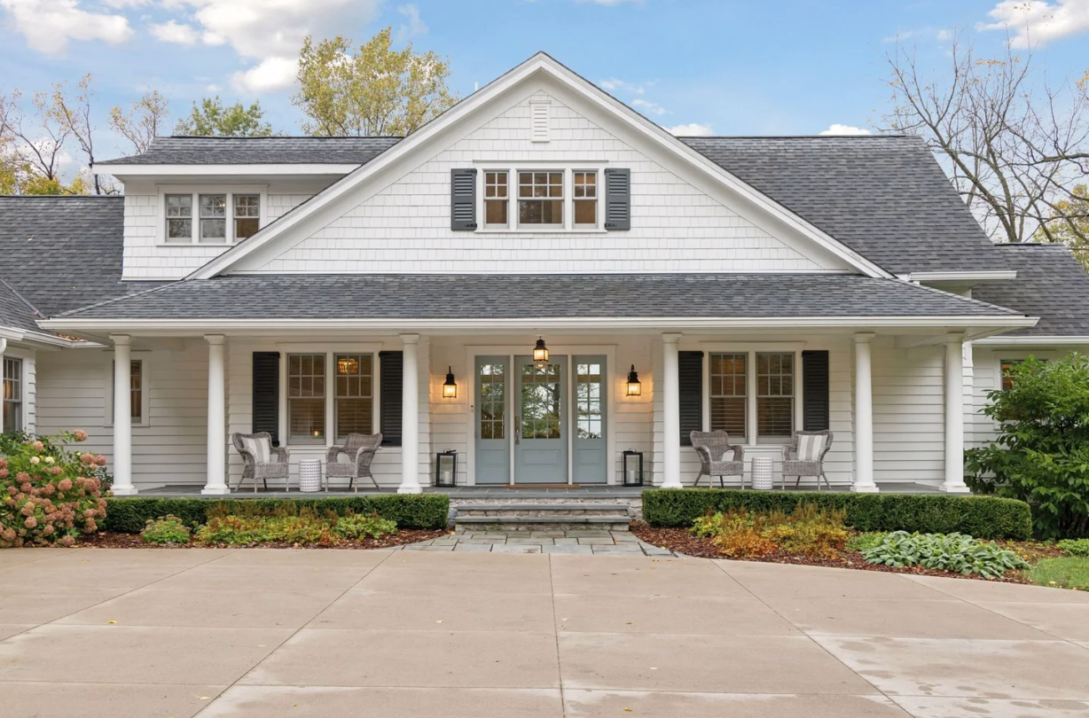 A white, two-story house with black shutters, a gray front door, and a front porch with seating and lamps, surrounded by landscaped bushes and trees under a blue sky.