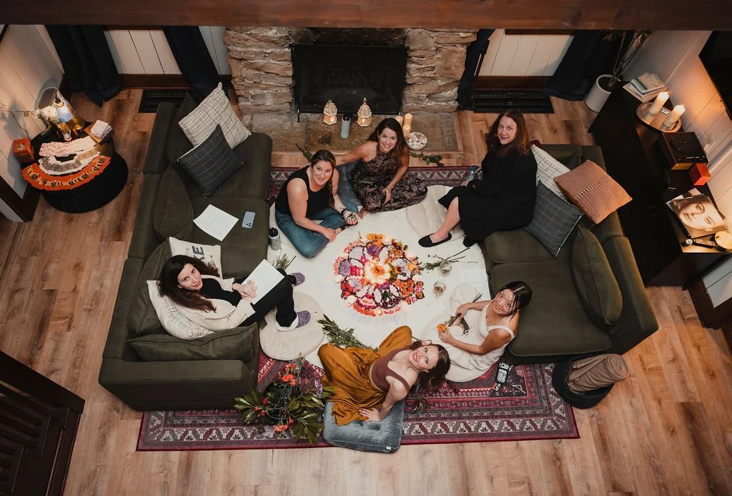 Five women sitting and lying on a carpeted living room floor around a floral centerpiece, with a stone fireplace and wooden flooring, cozy lighting, and various decorative items visible.
