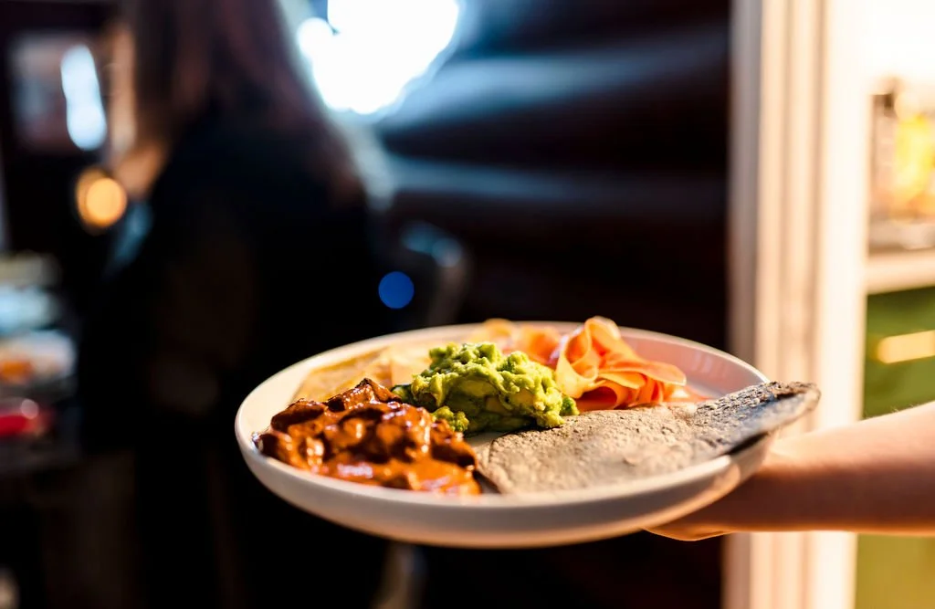 A plate of food held by a person's hand, featuring beans, guacamole, sliced turkey, and a fish fillet, in an indoor setting with a blurred background.
