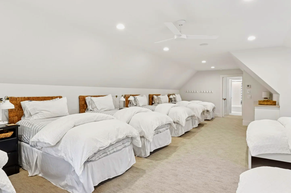 A spacious bedroom with white walls, multiple beds with white bedding and rattan headboards, ceiling fan, small nightstands with lamps, and a door leading to an outdoor space.