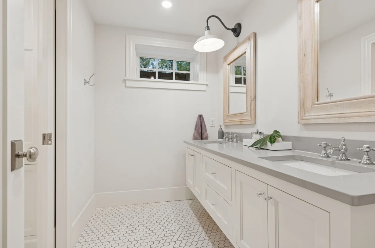 White bathroom vanity with double sinks, wooden framed mirrors, a wall-mounted light, and a small window with a view of trees.