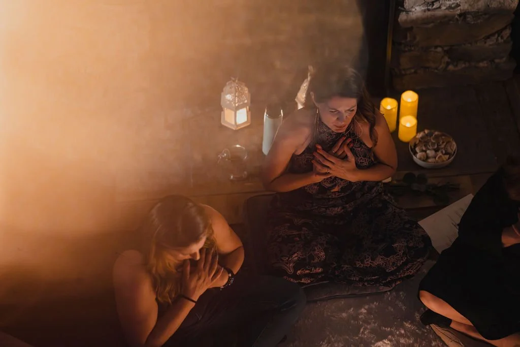 Three women praying with candles and a bowl of shells nearby, in a dimly lit room.