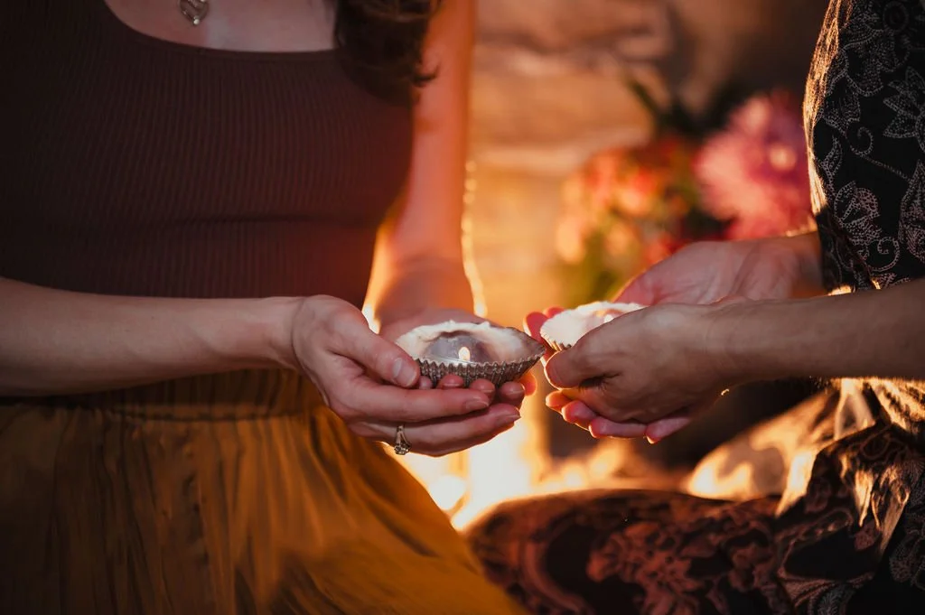 Two women exchanging cupcakes with lit candles. One woman wears a sleeveless top and the other wears a patterned sleeve. They are outdoors at sunset with flowers in the background.