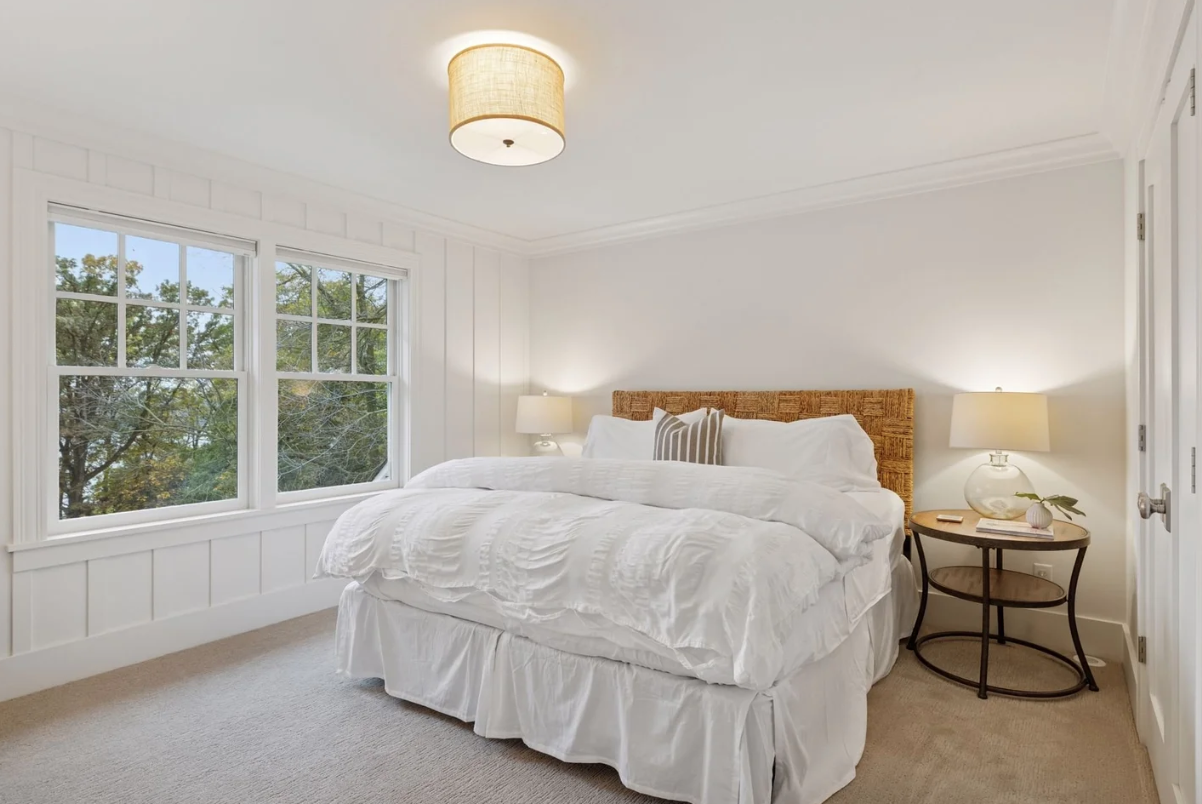 Bedroom with large window, white bedding, wicker headboard, bedside lamps, and a round wooden side table.