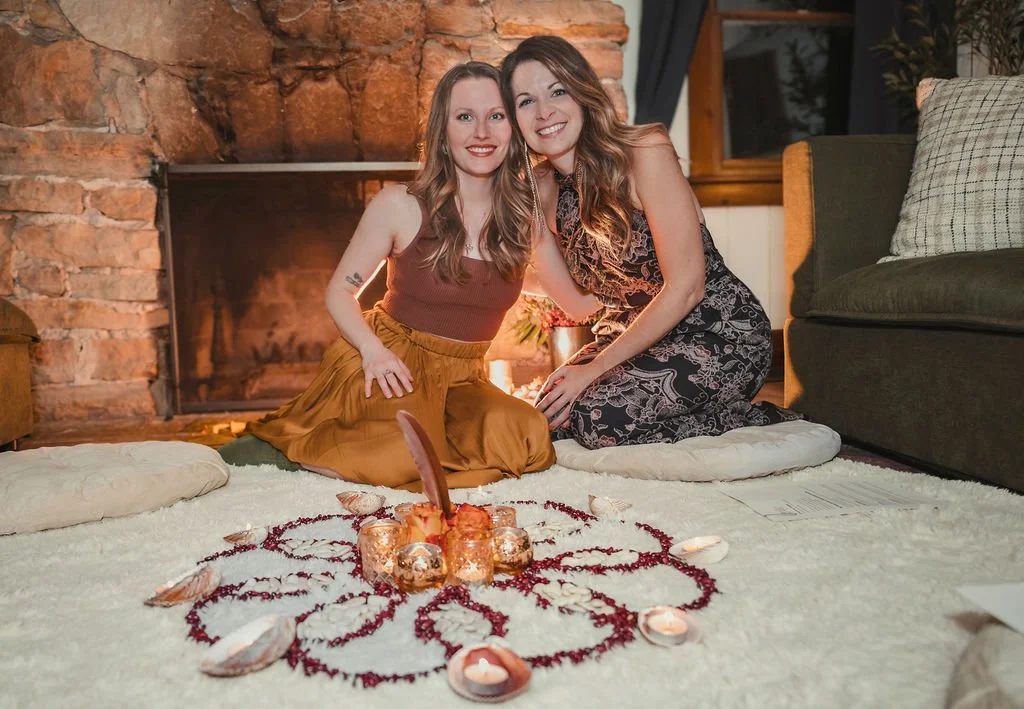 Two women sitting on a white rug in front of a brick fireplace. They are smiling and leaning toward each other. In front of them, there is a decorative arrangement of candles, flowers, and feathers, surrounded by red and white petals. One woman is wearing a brown sleeveless top and mustard-colored skirt, while the other is in a patterned dress.