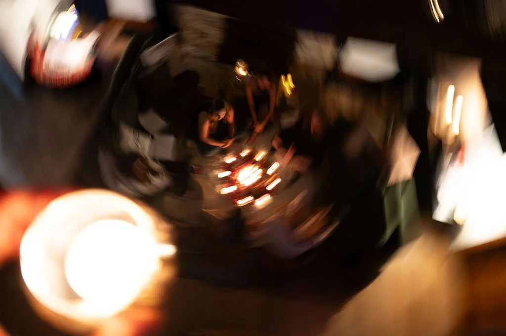 Blurred image of people gathered around a small fire in a dimly lit room, with a reflection seen on a shiny, curved surface.
