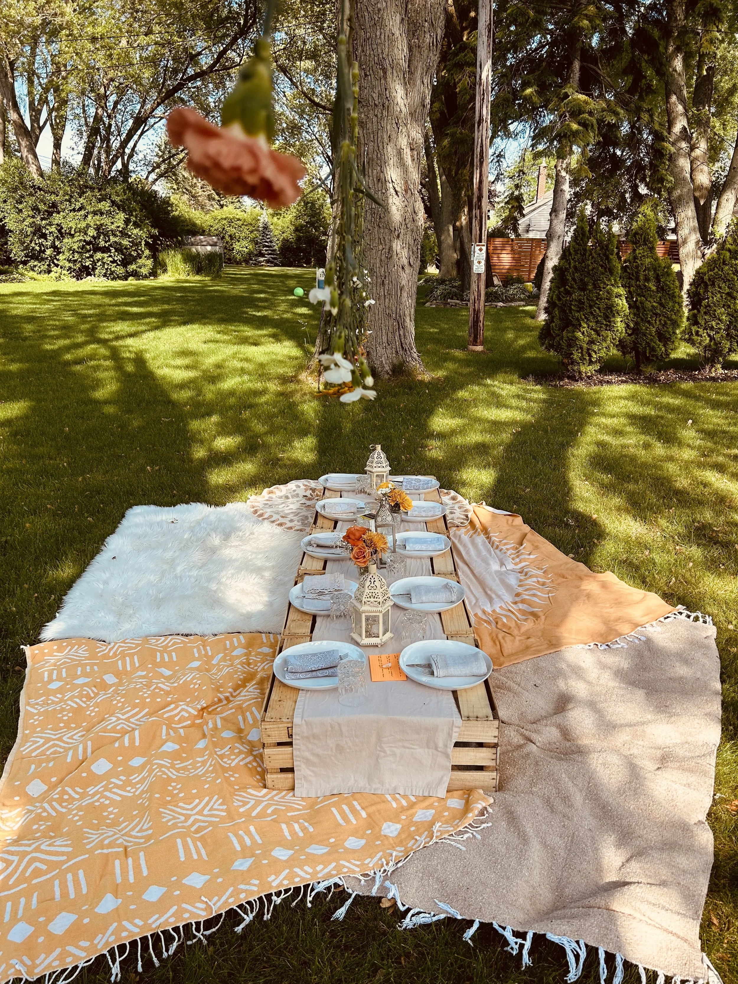 A daytime outdoor picnic setup with a low table on a yellow and white patterned blanket, surrounded by placemats, plates, glasses, and floral decorations, on a grassy lawn under trees.