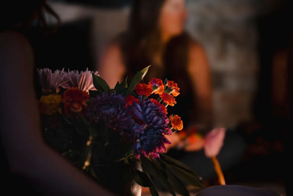 A bouquet of colorful flowers with a person sitting nearby, blurred in the background in dim lighting.