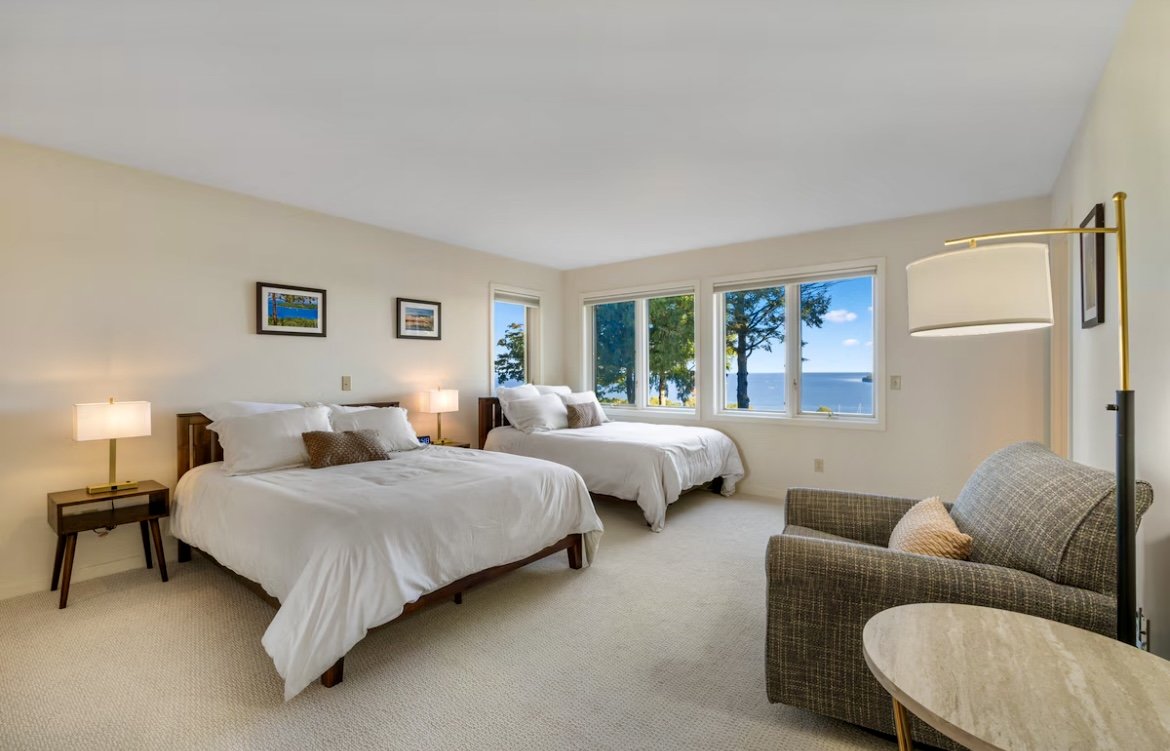Hotel room with two beds, a window view of trees and water, a sitting area with an armchair, side tables, and lamps.