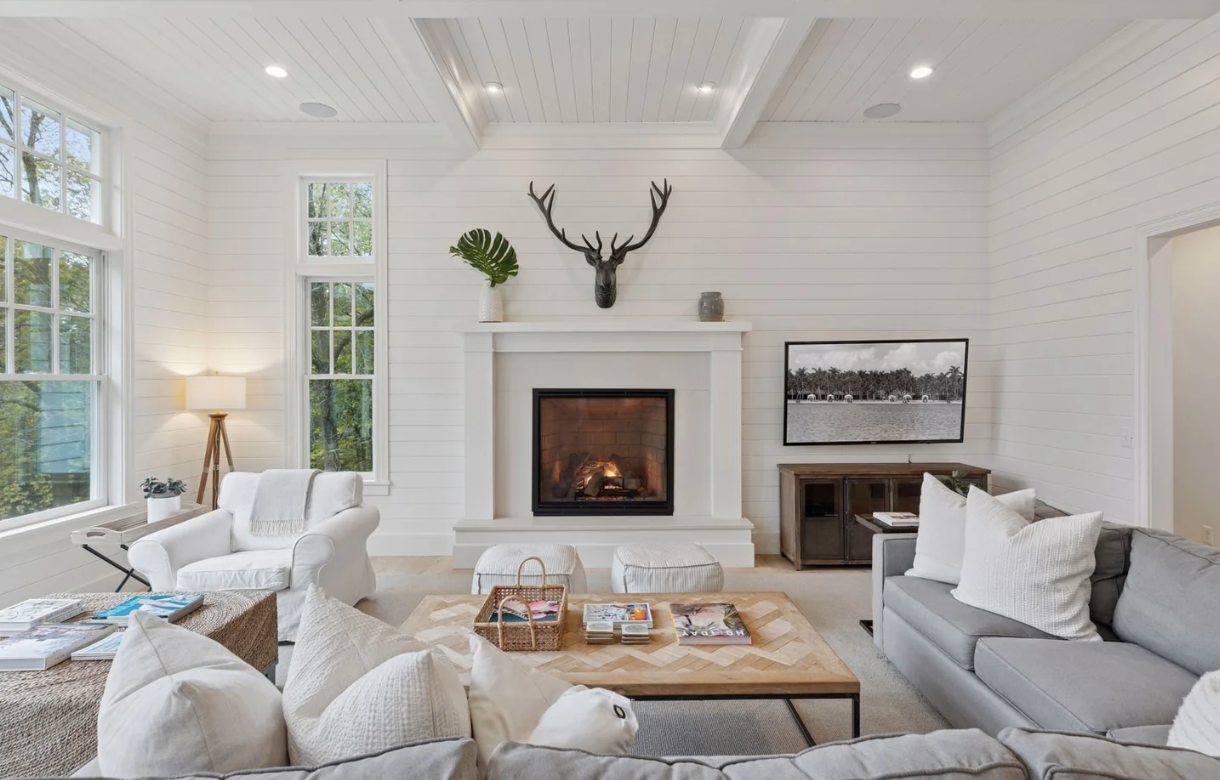 The beautiful environment for the Emerge spiritual wellness retreat. Living room with white walls, large windows, a fireplace, a mounted decor of a deer head, a flat-screen TV on a cabinet, and comfortable seating.