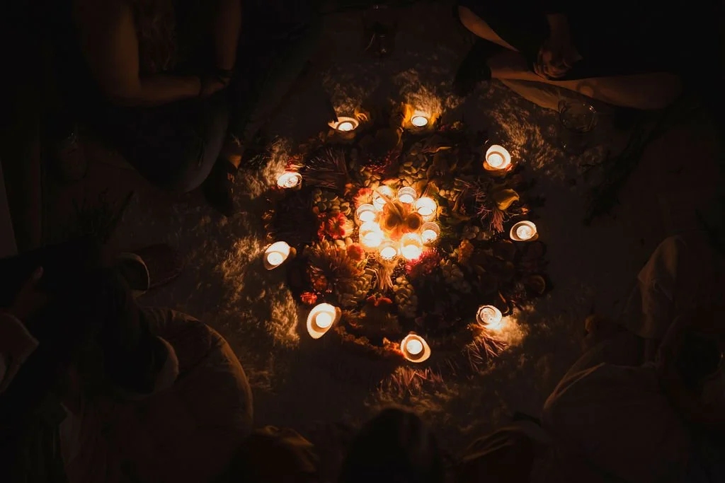 People sitting in a circle around a centerpiece of flowers and candles during a nighttime gathering or ceremony.