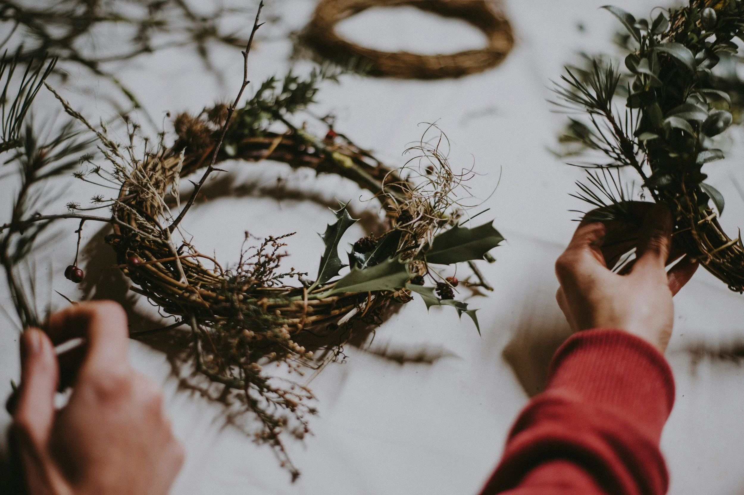 Two hands are shown making a wreath from various natural elements including twigs, holly leaves, berries, and pine needles, on a white surface.